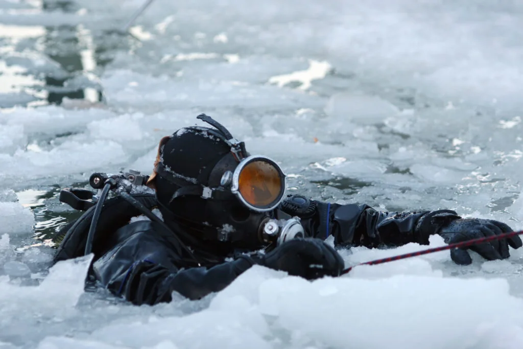Cold-water scuba diver wearing an insulated drysuit and hood surfacing through a hole cut in thick ice